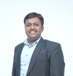 Professional headshot: man in a dark blazer and light blue shirt smiles at the camera against a light background.