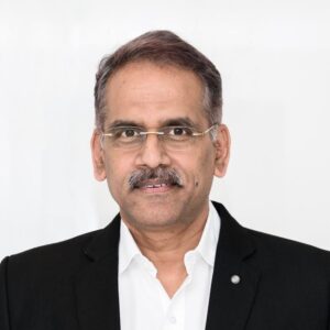 Portrait of a middle-aged man with gray hair and glasses, wearing a black blazer and white shirt against a light background.